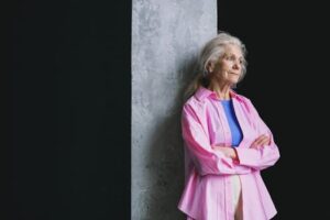 Senior woman with grey hair leaning against a wall, wearing a pink shirt, arms crossed, looking thoughtful.