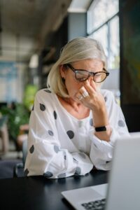 A senior woman in polka dot blouse appears stressed while using a laptop indoors.